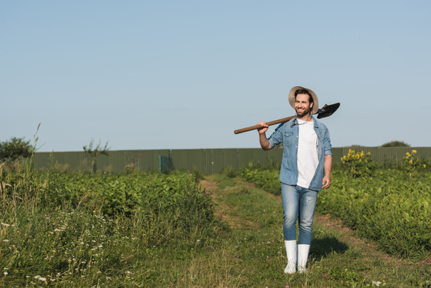 plný pohled na šťastného farmáře kráčejícího s lopatou na zeleném poli  - Fotografie, Obrázek