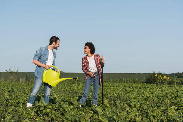 farmer with watering can talking to cheerful african american woman in field - Photo, Image