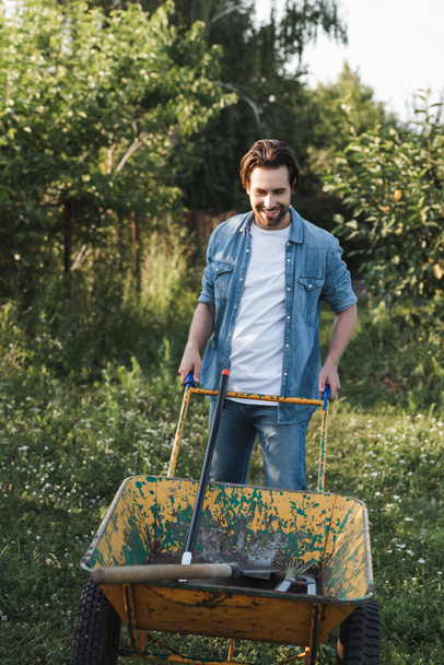 smiling farmer in denim clothes near wheelbarrow with gardening tools - 写真・画像