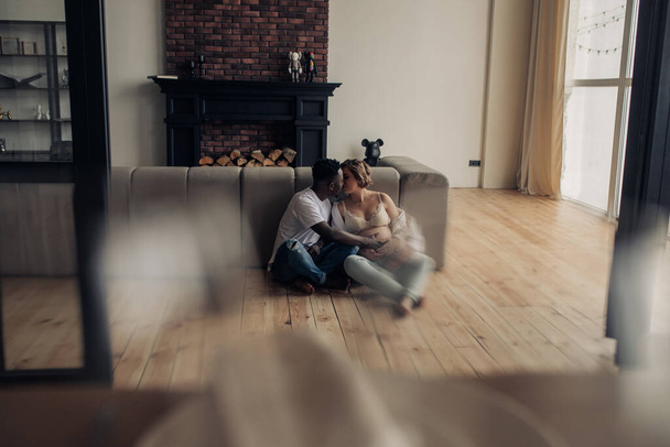 Jonge Afrikaanse man en zijn blanke zwangere vrouw zitten samen op de vloer naast de bank en omhelzen en kussen. Interraciaal huwelijksconcept. - Foto, afbeelding