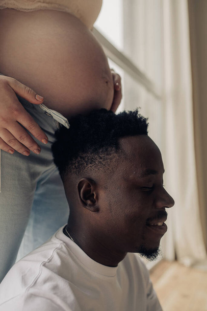 Vista de perto do rosto do homem africano feliz próxima barriga sua mulher grávida caucasiana. Conceito de casamento inter-racial. - Foto, Imagem