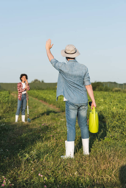 farmer with watering can waving hand to blurred african american woman in field - Foto, imagen