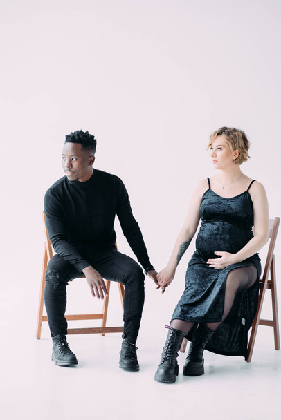 Young african man and his caucasian pregnant woman sit on chairs and hold hands with each other. Interracial marriage concept. - Fotoğraf, Görsel