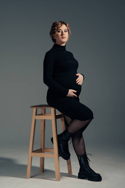 Young pregnant woman sits on chair against gray background. Maternity and pregnancy concept. - Photo, Image