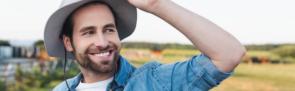положительный фермер в полях шляпы улыбаясь, глядя в сторону на открытом воздухе, баннер - Фото, изображение