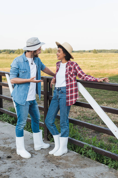 full length view of interracial farmers in hats talking near fence on farm - Photo, Image