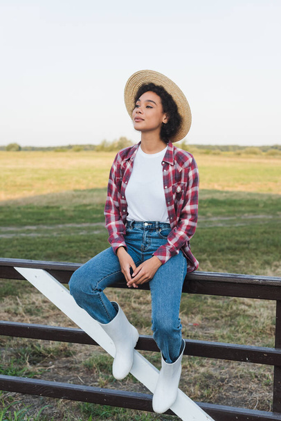 木製のフェンスの上に座っている再生シャツやジーンズでアフリカ系アメリカ人女性の完全な長さビュー - 写真・画像