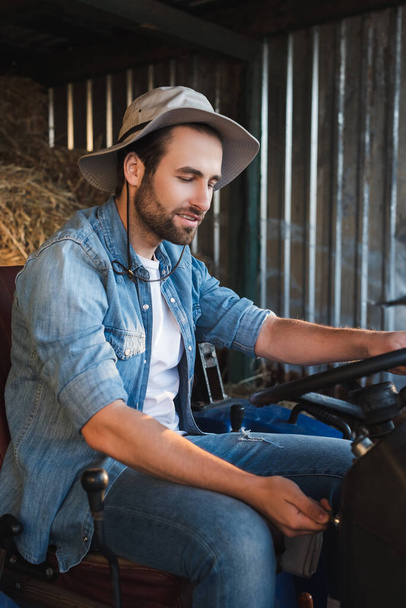 bearded farmer in brim hat and denim shirt sitting on tractor on farm - Фото, зображення