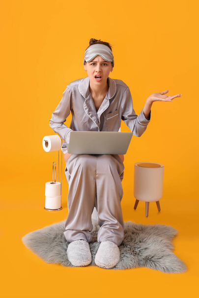 Δυσαρεστημένη νεαρή γυναίκα με πιτζάμες και φορητό υπολογιστή που κάθεται σε λεκάνη τουαλέτας με φόντο το χρώμα - Φωτογραφία, εικόνα