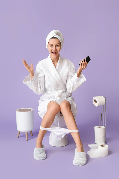 Happy young woman in bathrobe and with phone sitting on toilet bowl against color background - Photo, Image