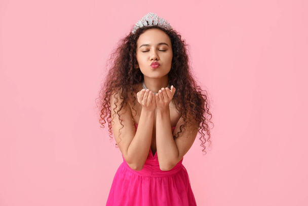 Beautiful young African-American woman in stylish dress and tiara blowing kiss on pink background - Photo, Image