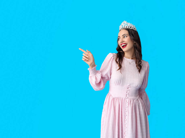 Beautiful young woman in stylish dress and tiara pointing at something on blue background - Photo, Image