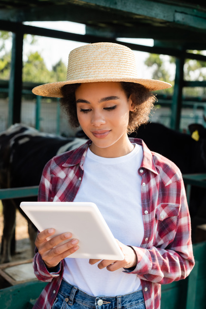 young african american woman in straw hat using digital tablet near blurred cowshed - Фото, изображение