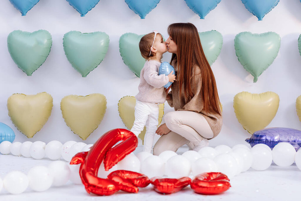 Красива молода мати і маленька мила дочка, розважаючись разом, стоять на барвистому фоні серцевих кульок, обіймаються і цілуються. день народження, оксамити, день матері
. - Фото, зображення