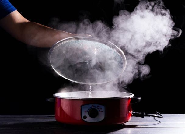 A séf keze kinyitja a forró leveses edény fedelét. füsttel fekete alapon az emberek főzés és forrásban lévő víz a konyhában - Fotó, kép