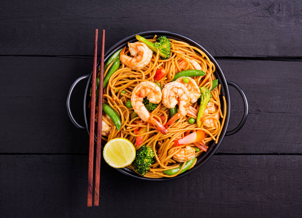 Жареные спагетти или жареная лапша с овощами и креветками в черной миске. темный фон, вид сверху - Фото, изображение