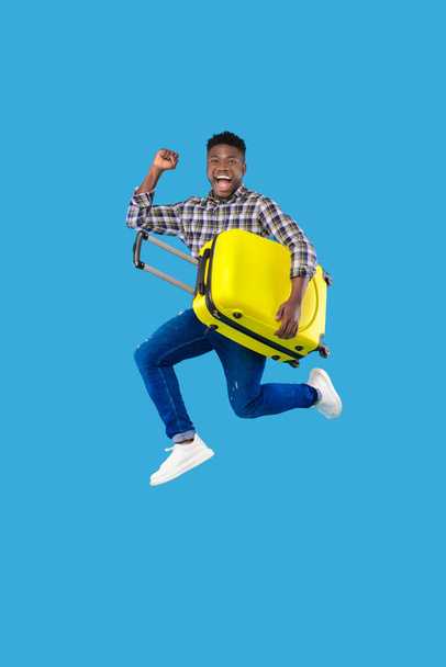 Jeune homme noir excité avec une valise lumineuse sautant et faisant un geste OUI sur fond bleu studio, espace de copie - Photo, image