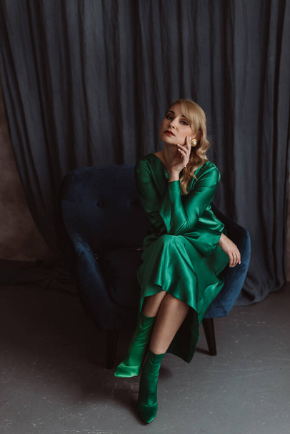 A beautiful blonde woman in an elegant green silk dress sits on a blue armchair against a dark background. Beauty industry, preparing for an important event. Soft selective focus. - Fotoğraf, Görsel