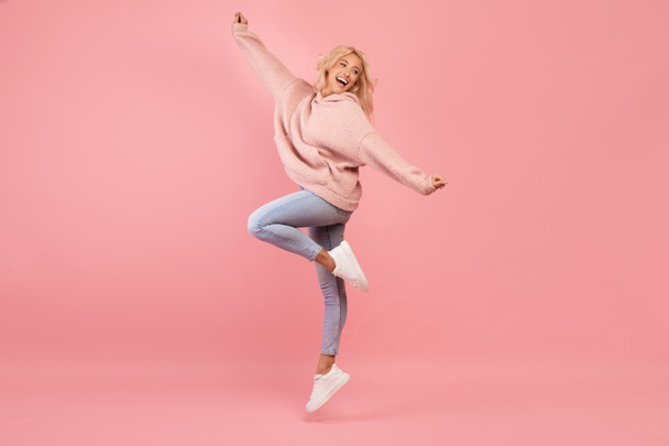 Χαρούμενη νεαρή κοπέλα που πηδάει, διασκεδάζει και χαμογελάει, είναι σε καλή διάθεση και χαζολογάει πάνω από ροζ φόντο - Φωτογραφία, εικόνα