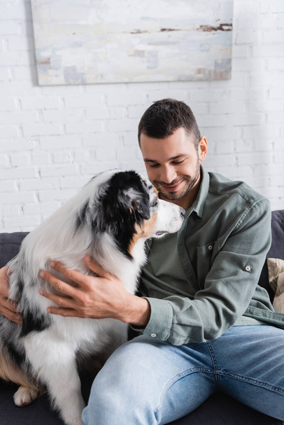 smiling bearded man cuddling australian shepherd dog on couch - Photo, Image