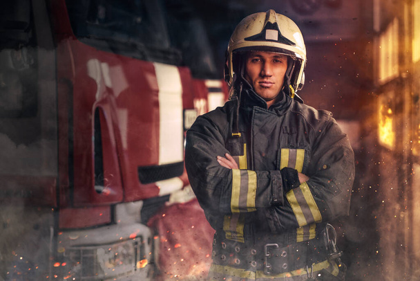 Храбрый пожарный позирует вокруг пожарной машины в помещении пожарной станции - Фото, изображение