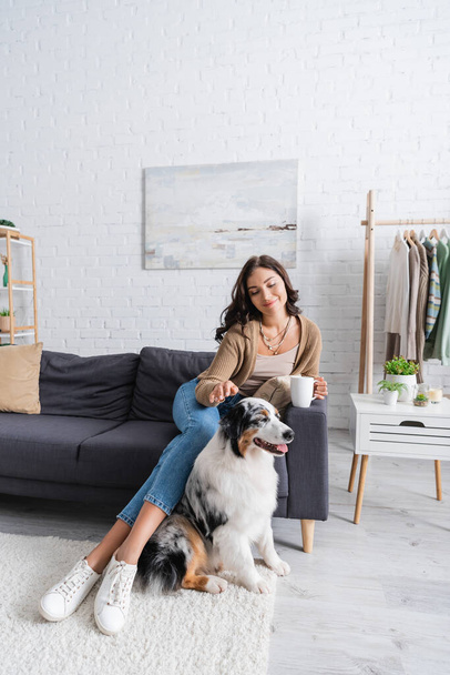 glückliche junge Frau sitzt auf der Couch und knuddelt australischen Schäferhund, während sie eine Tasse Kaffee hält - Foto, Bild