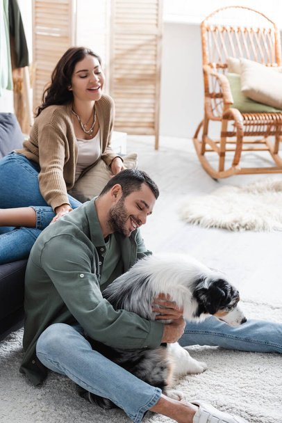 щасливий бородатий чоловік обіймає австралійську вівчарку біля радісної дівчини на дивані
 - Фото, зображення