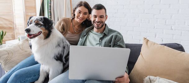 счастливая молодая пара смотрит кино на ноутбуке возле австралийской овчарки, баннер - Фото, изображение