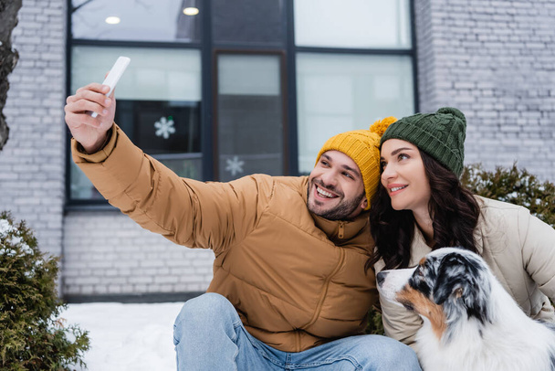 felice uomo in maglia cappello prendendo selfie con fidanzata e cane pastore australiano in inverno  - Foto, immagini