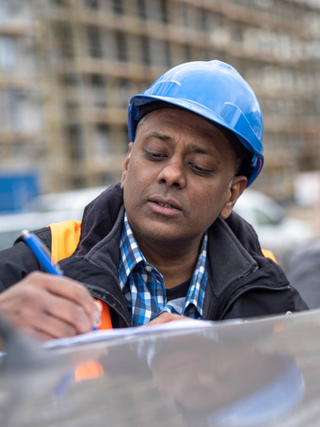 Ingénieur indien portant des vêtements de travail de protection vérifiant ses projets et signant des documents, à l'extérieur - Photo, image