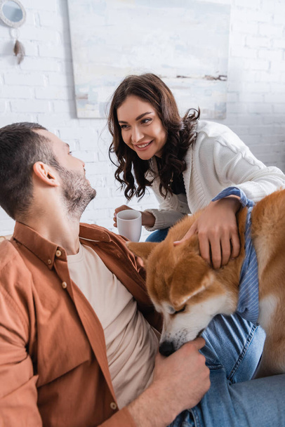 Bir fincan kahve tutan mutlu kadın ve Akia Inu köpeğinin yanındaki sakallı adama bakıyor. - Fotoğraf, Görsel
