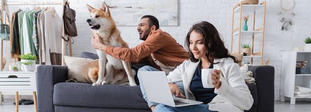 Nuori nainen kupin avulla kannettava tietokone lähellä poikaystävä halailu akita inu koira sohvalla, banneri - Valokuva, kuva