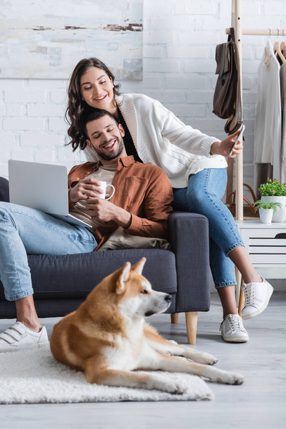 smiling young woman holding smartphone near boyfriend with laptop and looking at akita inu dog in living room - Photo, Image