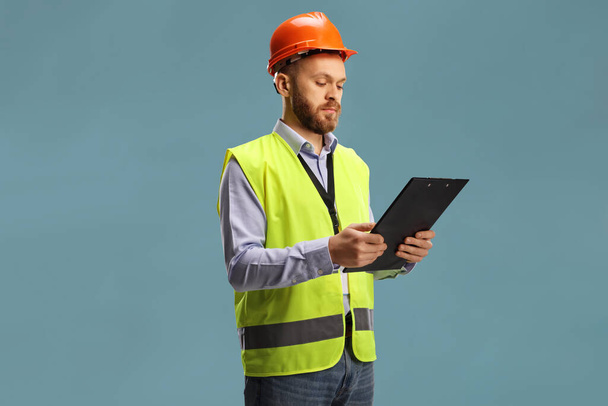 Male engineer wearing a reflective vest and helmet holding a clipboard isolated on blue background - Foto, Bild