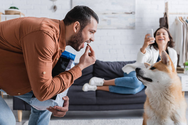 bearded man holding pet food near open mouth and looking at akita inu dog near blurred girlfriend with smartphone - Photo, Image