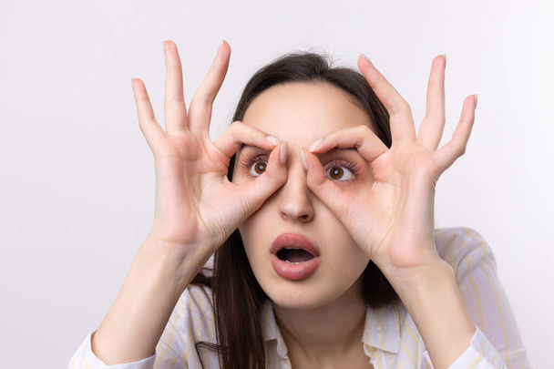 Close up portrait of attractive quirky young woman making binoculars with hands showing ok gesture on white studio background. - Фото, зображення