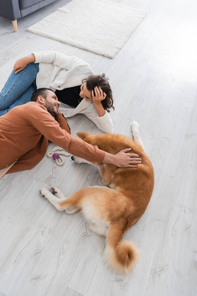 top view of joyful young couple looking at each other while lying of floor with akita inu dog - Photo, Image