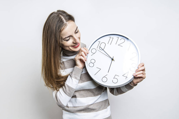 Mujer joven sonriente mirando el reloj de pared blanco sobre fondo claro  - Foto, imagen