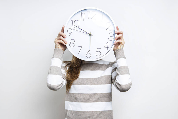 Giovane donna che tiene un orologio da parete che si copre il viso su uno sfondo leggero  - Foto, immagini