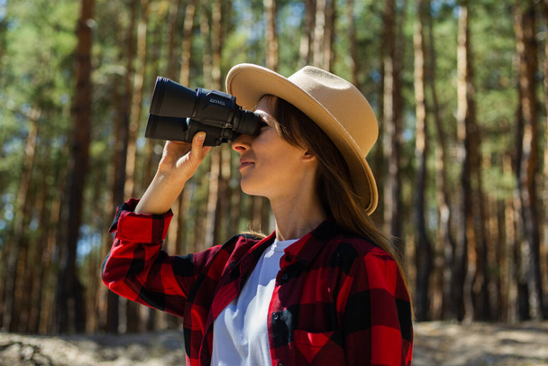 Woman in hat and red plaid shirt looking through binoculars in the forest. - Foto, Imagen