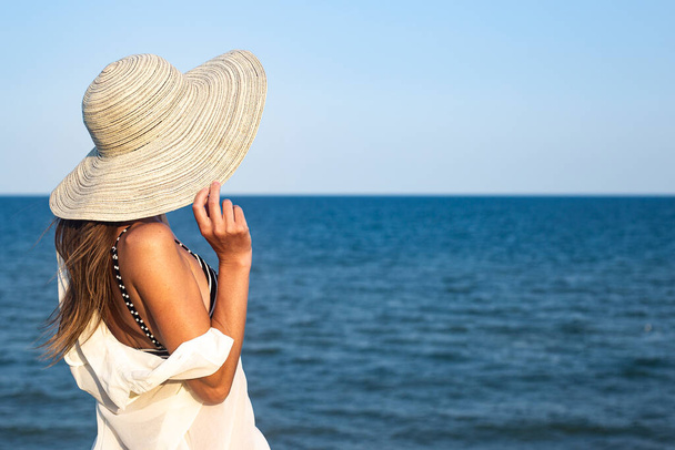 Νεαρή γυναίκα με καπέλο σε γκρεμό κοιτάζει τη θάλασσα μια καλοκαιρινή μέρα  - Φωτογραφία, εικόνα