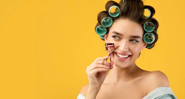 Happy Woman Using Hair And Eyelashes Curlers Over Yellow Background - Foto, imagen