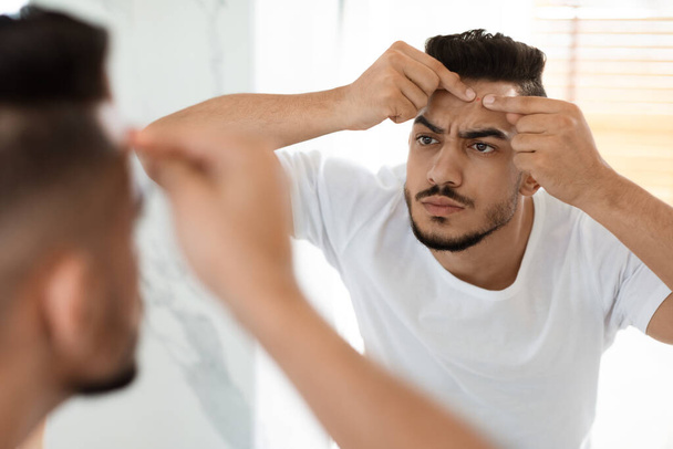 Vertoornd Arabisch man met probleem huid popping puist op voorhoofd in de buurt van spiegel - Foto, afbeelding