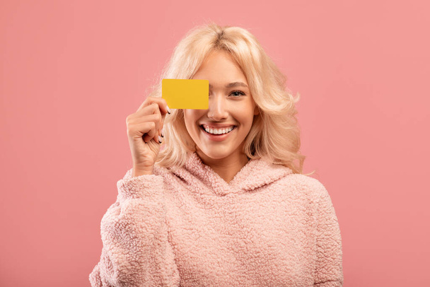 Kauf, Geld und Zahlungskonzept. Hübsche Dame bedeckt Auge mit Kreditkarte, posiert auf rosa Wand Hintergrund - Foto, Bild