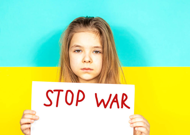 Ένα κορίτσι κλαίει, κρατώντας μια αφίσα STOP WAR στα χέρια της στο φόντο της ουκρανικής σημαίας. Σταματήστε τον πόλεμο στην Ουκρανία. Η έννοια της ειρήνης στην Ουκρανία. Τόπος για κείμενο επιγραφής - Φωτογραφία, εικόνα