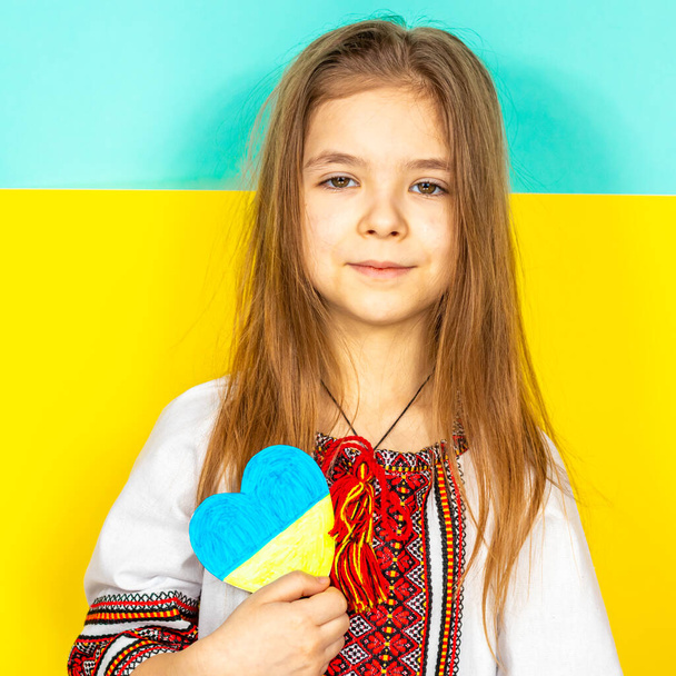 Ein Mädchen in einer nationalen ukrainischen Wyschywanka zeigt ein Herz in gelben und blauen Tönen als Zeichen der Liebe zur Ukraine, in Großaufnahme vor dem Hintergrund der ukrainischen Flagge. Das Konzept des Friedens in - Foto, Bild