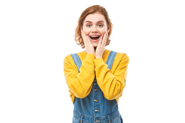 Shocked face red-haired girl looks surprised with open mouth holding head with hands on white studio background - Foto, imagen