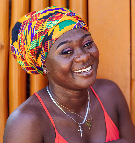 カラフルな頭を持つアフリカの女性と希望に満ちた笑顔は、西アフリカのケタ・ガーナの熱帯の村で描かれた木製の壁に向かって立っています - 写真・画像