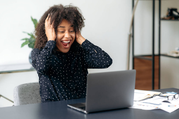 Enthusiastic joyful successful smart emotional dark-skinned business woman, broker, freelancer, sitting in office with a laptop, happy for a good deal, victory, holding her head and shouting joyfully - Fotó, kép
