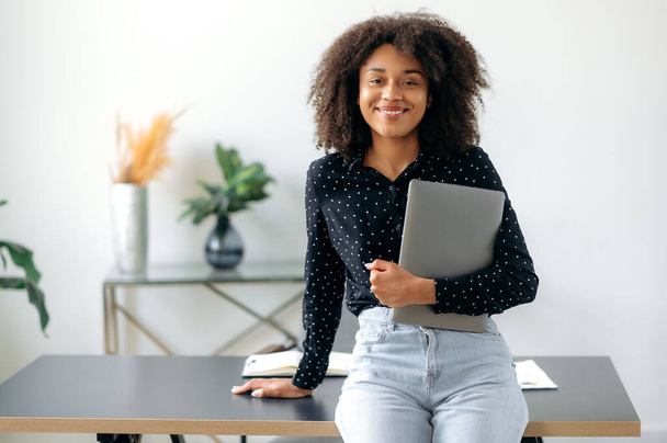 African American pretty, curly haired, confident young adult business woman or manager standing near work desk in office, trendy dressed, holding laptop, looking at camera, smiling friendly - Photo, Image